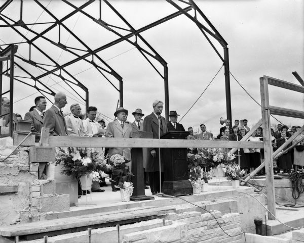 Speakers stand near the front of the partially finished new building during the cornerstone laying ceremony at Trinity Lutheran Church, 1904 Winnebago Street at First Street. At the speakers podium is Herman Ekern, president of the Lutheran Brotherhood. Officials in back of him, left to right, are: Alvin Nygaard, president of the Trinity congregation; Rev. J.N. Walstead, pastor emeritus at Trinity; Rev. Enoch Tetlie, president of the Madison circuit of the Evangelical Lutheran Church; Rev. Robert G. Borgwardt, pastor at Trinity; Governor Oscar Rennebohm; Rev. F.I. Schmidt, pastor at Bethel Lutheran Church; and City Councilman Herbert C. Schenk.