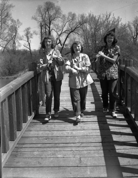 Three teenage girls are carrying shovels on their shoulders while crossing a wooden foot bridge.  More than 150 Madison high school pupils recruited through the Madison Youth Council helped plant trees and shrubs on the Vilas Park island.