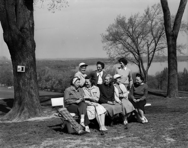 Women Golfers gather around a bench at Blackhawk County Club on Ladies' Day. Seated from left to right are: Mrs. A.J. McAndrews, Betty Geisler, Nettie Peterson, Florence Burdick, and Valerie Coan. Standing left to right: Bess Smith, Jessie Morgan, Isabel Peterson. Lake Mendota is visible in the background.