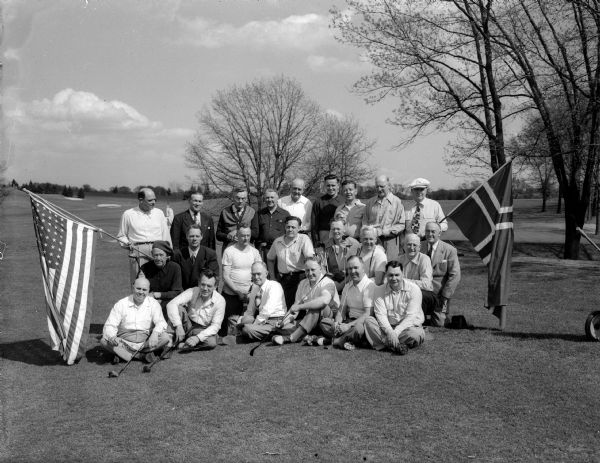 Twenty-three men, participants at the Ygdrssil Literary Society's Syttende Mai golf tournament and dinner, posing between an American flag and a Norwegian flag at Nakoma Golf Course. The Ygdrssil Literary Society was a Norwegian language and culture club.