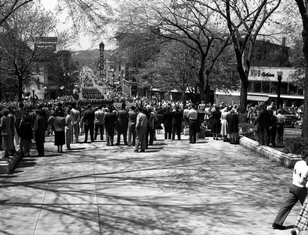 Spectators are shown standing on the steps at the Capitol Square looking down State street at approaching U.W. ROTC marching units. The view includes the entire length of State street and Bascom hill. The parade commemorates Armed Forces Day.