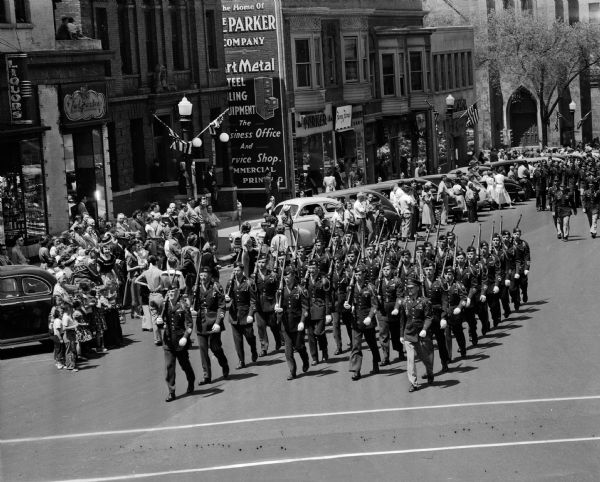 University of Wisconsin ROTC marching units moving south down South Carroll street during a parade commemorating Armed Forces Day. The Episcopal cathedral is in the upper right hand corner. Spectators are watching from the sidewalk and in the street between diagonally parked cars.