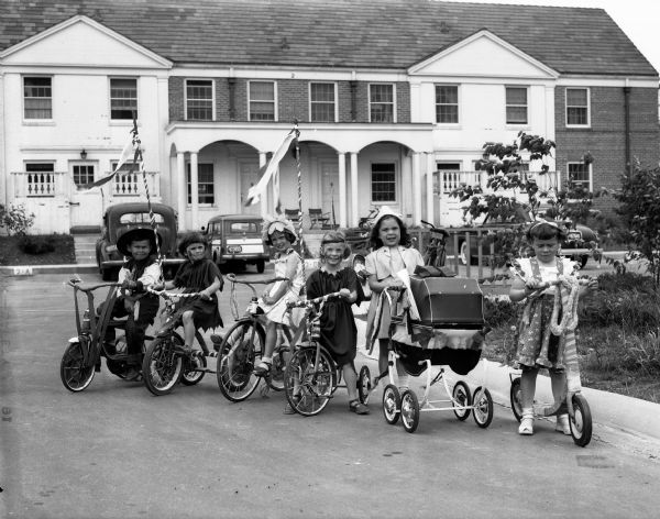 Children of University of Wisconsin faculty living in University Houses at Eagle Heights are costumed as Native Americans, cowboys, and flowers are marching in a Memorial Day parade on gaily decorated tricycles. Pictured left to right are: Eddie Geaney, Mary Denise Luckhardt, Marilyn Weinburg, Deborah Lea Luckhardt, Jeanne Busacoa, and Adrienne Arnold.