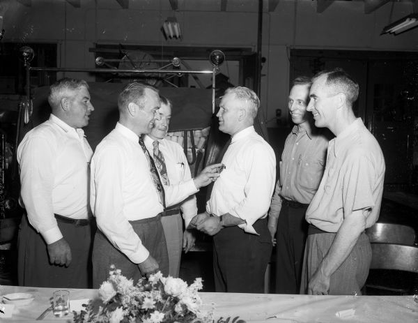 Six firefighters are shown at First Assistant Fire Chief Leonard Sime's retirement party inside the Central Fire Station, 18 South Webster Street. Lieutenant Wilson H. Donkle (second from left) president of the Madison Firefighters Local No. 311, is shown presenting an honorary badge on behalf of the union to Sime (fourth from left). Pictured left to right are: Assistant Chief Edward Durkin, who succeeds Sime as first assistant chief; Donkle; Fire Chief Edward Page; Sime; Edward Bonkins, who served as master of ceremonies and Capt. William Lynaugh who will become second assistant fire chief.
