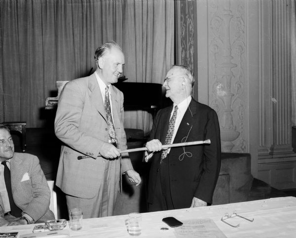 Carl Runge (right), 91, U.W. Madison 1886 alumni and a former Milwaukee judge, is shown receiving a gold top cane to honor his being the oldest alumnus in attendance at the Half Century club luncheon at the Great Hall at Memorial Union. The cane is being presented by John Berge (left), executive secretary of the Wisconsin Alumni association.