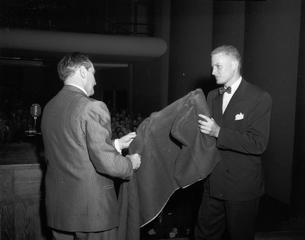 Two men, one of which is possibly Manchester Morgan, presenting or receiving what appears to be a blanket in Memorial Union Theater(?).