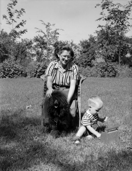 Marcella Candlin, active member of the Badger Kennel Club, and her miniature French poodle, Colette, are shown sitting outside. At their feet, playing with a toy cash register, is her son, Bruce Candlin.