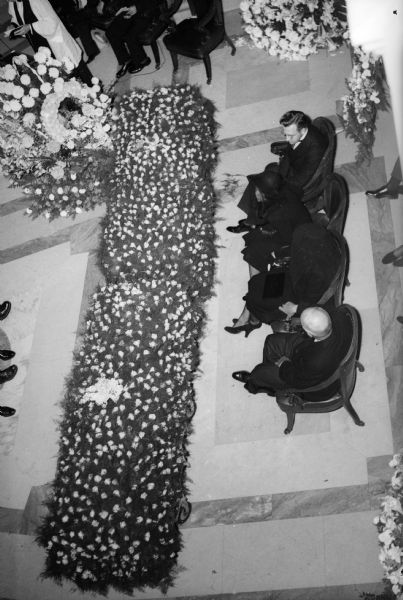 Elevated view of the twin caskets of University of Wisconsin president Glenn Frank and Glenn, Jr. in the Wisconsin State Capitol rotunda at the state funeral services. Seated in front of the caskets are Tom K. Smith (foreground), Mrs. Frank's brother; Mrs. Frank; Mary Jane Manierre, the woman Glenn, Jr. was to marry; and Roy L. Matson. The father and son died September 15, 1940 in an automobile accident south of Green Bay while campaigning for the Republican nomination for the United States Senate.