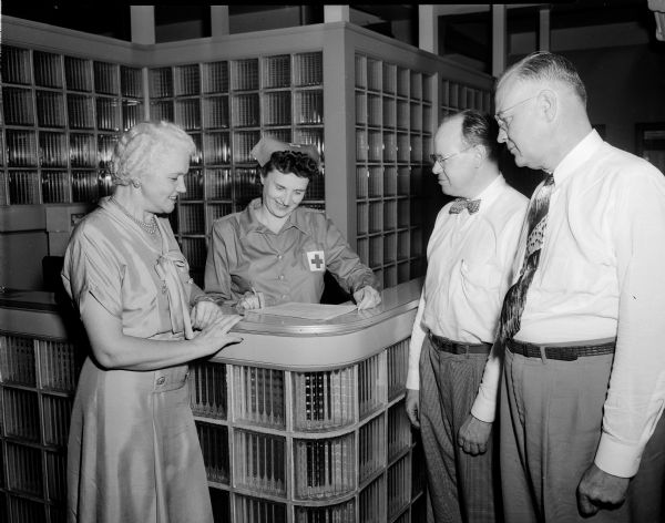 Mrs. Oscar (Mary) Rennebohm is shown registering at the American Red Cross regional blood center in Madison. Mrs. Charles Schmalbach, volunteer officer of the day, is shown greeting and checking in Mrs. Rennebohm and two other donors, John Canfiels (left), and Alan Hackworthy (right), both blood program committee members.