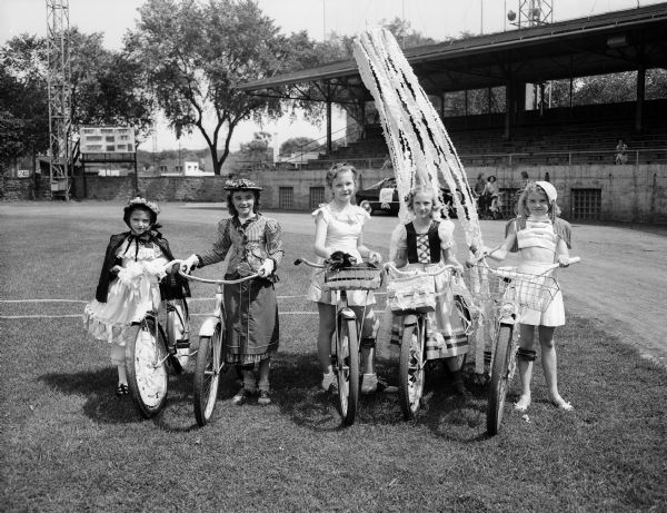 Girl costume winners in the Montgomery Ward Bicycle Safety Parade pose for a portrait with their bikes on a football field. Pictured left to right are: Mary Kay Sullivan, Kay Keliher, Judith Johnson, Sharon Miller, and Mary Lou Carlson.
