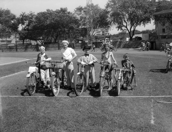 Boy costume winners in the Montgomery Ward Bicycle Safety Parade pose for a portrait with their bikes on a football field.  Pictured left to right are: Jon Buschke, James Stewart, Jackie Shipler, William Miller, and Billie Dunkel.