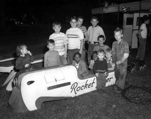 Children who came to the UNICO festival in Brittingham Park gather around a soap box derby car for a group portrait. The festival is sponsored by the Madison chapter of UNICO, which stands for Unity, Neighborliness, Integrity, Charity, and Opportunity.