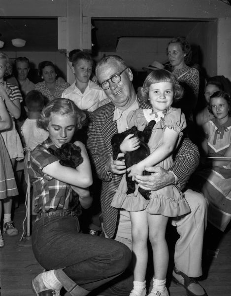 The Badger Kennel Club sponsored an essay contest on "Why I Should Own A Dog" and awarded purebred Cocker Spaniel puppies to winner Janice Emanuel and runner-up Gloria Blindheim. The girls are shown holding their new puppies as <i>Wisconsin State Journal</i> columnist "Roundy" Coughlin is looking on.
