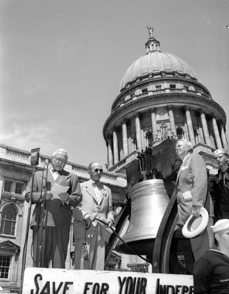 Governor Rennebohm delivers an acceptance speech after receiving a liberty bell replica from Vern Bell.  The men are, from left to right, Governor Rennebohm, Vern S. Bell, Chairman of Dane County U. S. savings bond committee, Harold F. Dickens of Milwaukee and State Director of the bond drive, and Franz Turner of Ship No. 501, Madison, Wis.