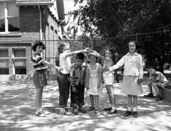 Six children engage in folk dancing at Randall School playground on Regent Street, while one bystander observes from the background. Pictured dancing from left to right are: Judy Wyngaard, Marsha Rasmussen, Dan Mermin, Mary Farrell, and Bellsy Oelschlager.