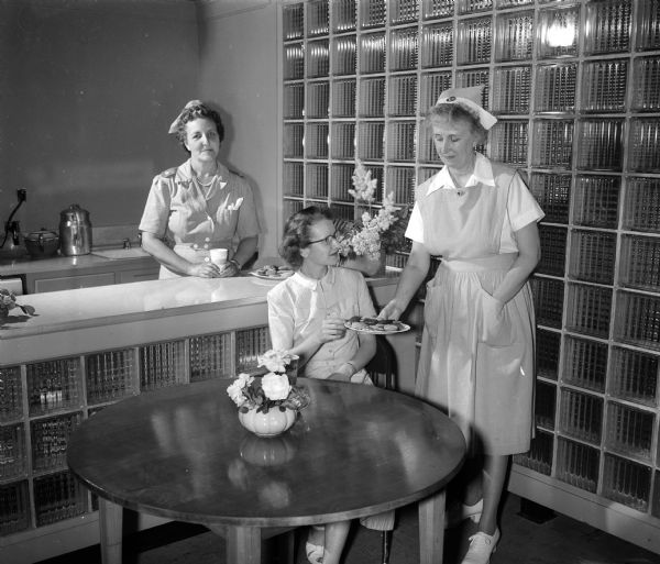 Volunteer worker Mrs. Charles (Elnora) Wirth stands at the counter in the American Red Cross Blood Center canteen, while nurses' aide Mrs. Gordon (Mabel) Nelson offers a snack to Grace Cuff, a blood donor.