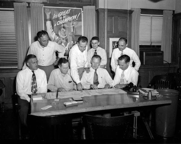 Organizers of the 1950 Madison Soap Box Derby gather around a table while examining papers. Pictured left to right, front row, are: Glenn "Pat" Holmes, John G. Thompson, Walther Thompson and James G. Marshall; left to right, rear row: Pete Norg, Don F. Knowles, Martin Wolman and Ted Bast. A Soap Box Derby poster is on the wall behind them.