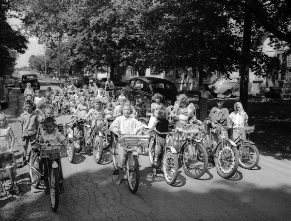 A large group of children with bicycles pose in the street in front of the Gallagher Plat playground.  In front are, left to right, are Karen Vanderbloemen, Gary Wesley Bakken, Carol Baun, and Mary Sime.