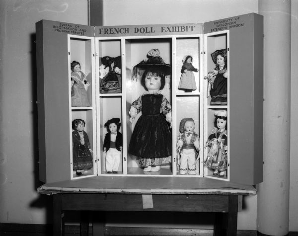 Nine dolls dressed in costumes of provincial France, donated by their French owners in gratitude to the people of Wisconsin for their contribution to the Friendship train of 1947, are arranged on display. The large doll in the center is from Burgundy. The others, from left to right in the top row, are from Arles of Provence; Alsace Loriane, Provence, and Loraine, and on the bottom row, Savoy, Provence, Basque, and Brittany.  The University of Wisconsin Extension Bureau of Information and Program Services to whom the dolls have been entrusted will send the collection out for display to interested Wisconsin residents.