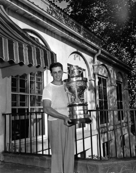 Mike Bencriscutto, Racine, standing in front of the Maple Bluff Country Club clubhouse while holding a large trophy.  He was the winner of the Wisconsin State Golf Association's Men's State Amateur golf tournament.