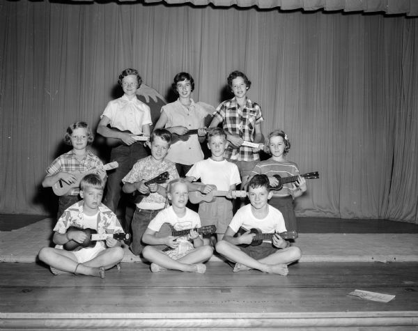 Group portrait of participants in the ukulele concert that was part of the "open house" of the Shorewood Hills summer children's recreation program. Pictured left to right are the children, taught ukelele skills by Shorewood Recreational director Richard Miyagawa: John Marshall Nelson, Robert Arnold, and Dick Meloy, in the front row; Marilyn Metz, Fritz Holmquist, Craig Washa, and Sharon Mack, second row; and Pricilla Butts, Judy Hicks, and Shirley Metz, top row.