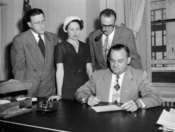 Acting Madison City Manager George Forster signs a proclamation designating the week of August 14 as Voters' Registration week.  Watching are, left to right: Al W. Bareis, city clerk, Mrs Arthur (Julia) Miles, chair of the "Register and Vote" committee, and Homs A. Schwahn, chairman of publicity and promotion for the committee.