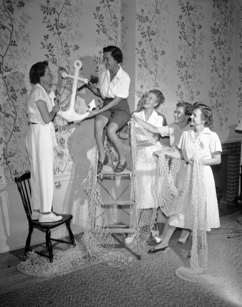 Members of the social committee for the Inland Lakes Regatta prepare the decorations at the Maple Bluff Country Club for some of the parties to be held during the regatta. The committee chairman, Mrs. Ray Footh, sits on the ladder. Other committee members are, left to right, Mrs. Gilman G. Page, Mrs. Timothy Brown, Mrs. Deane Adams, and Mrs. Walter Stebbins.