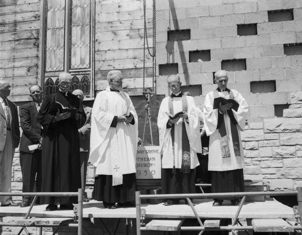 Men stand on a platform during the cornerstone-laying dedication ceremony for the reconstructed Norway Grove Lutheran church. Taking place in the ceremony, left to right are: Alvin Neuman, president of the congregation; the Rev. Peter Johnson, DeForest, retired pastor; Simon Ethun, chairman of the building committee; the Rev. O. J. H. Preuss, a former church pastor;  the Rev. Axel Anderson, church pastor; and the Rev. C. G. Naeseth, pastor of the Spring Prairie Lutheran church.