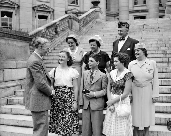 Secretary of State Fred Zimmerman, left, greets three war orphans visiting Madison under the auspices of the Veterans of Foreign Wars auxiliary. Left to Right: Margaret Craton, 17, England; Konstantinos Sotirhof, 15, Greece; and Beatrice Li Donnici, 17, Italy. The adults in the background include Mrs. Gwendolyn Johnson, Richland Center, youth activities chairman of the state VFW auxilary; Mrs. Rose Schell, Milwaukee, department president of the VFW auxilary; Lyall T. Beggs, Madison, former national commander-in-chief of the VFW; and Miss Josephine Brahm, president of the Madison VFW auxiliary.