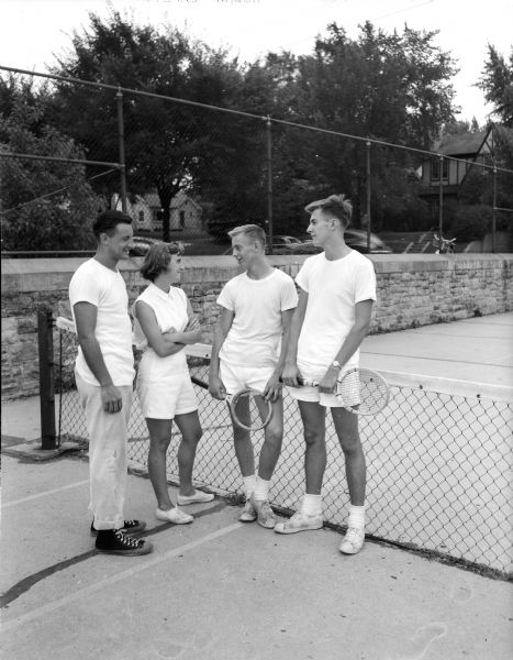 Four tennis tournament enthusiasts are shown with racquets in front of a net and stone wall. They are coach Robert Brill, Helen MacDuffee, Don Peterson and Owen Roberts.
