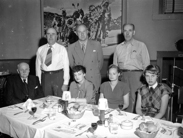 Three men stand behind a group of men and women seated at a banquet table in the Indian Room at the Monona Hotel, for a Gunderson-Finn party.