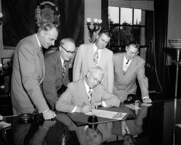 Governor Oscar Rennebohm, seated, signs a proclamation that officially opens National Home Week in Wisconsin. With him at the signing, standing left to right: Frank W. Cortwright, Washington, D.C., executive vice-president of the National Home Builders Association (NAHB); E.B. Sokoloski, president, Madison Builders Association; John McKenna, publicity chairman for the Madison group; and Lowell Gerretson, executive director of the Wisconsin and Madison Builders associations.
