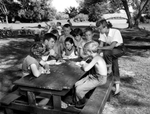 A group of kids play a paper game "Air Defense" with two playground directors at a table in Vilas Park. Sitting around the table, from left, are: Nancy Brinkman, director Darrell Dillman, John Drake, Calvin Laws, Naomi Shapiro, Jane Debs, Gordon Kraut, Jerry Doran, and director Bev Melvin.