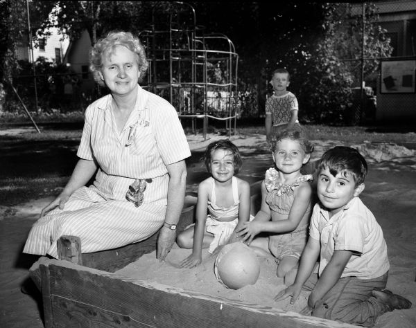 Mrs. Irene M. Borman, playground director at the Milton-Charter park for twelve years, sitting at a sand box with youngsters Joane Bruno, Kay Kerwin, and Tony Balsamo.