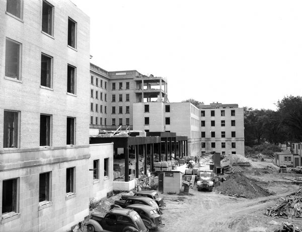 Wisconsin General hospital, located at 1300 University Avenue, is shown during the construction of the front and double wing expansion. The framework for the new entrance to the building is in the center. There are four parked cars, a work truck and construction rubble along the exterior of the building.