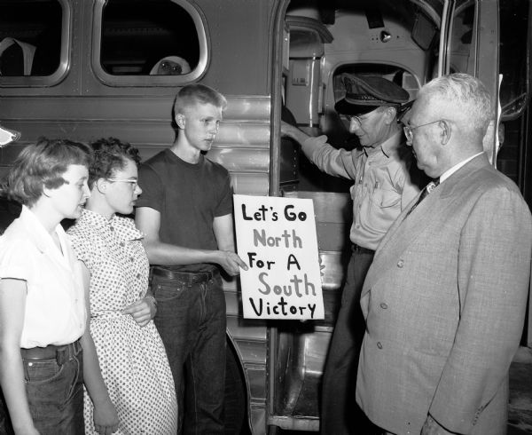 Three students and two adults are shown making arrangements for the chartering of a bus to take Madison high school students on a chaperoned trip to Green Bay to the fifth annual North-South All-Star high school football game. Shown left to right standing by a bus door are Phyllis Gilbert, Joan Gilbert, and Youth Mayor John Novotny holding a sign stating: " Let's Go North For A South Victory" and Ervin Kuhnz, bus driver, and Louis A. Leonard, bus company superintendent.