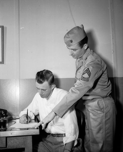 A Marine corps officer stands behind a seated recruit in the reserves as he fills out papers.