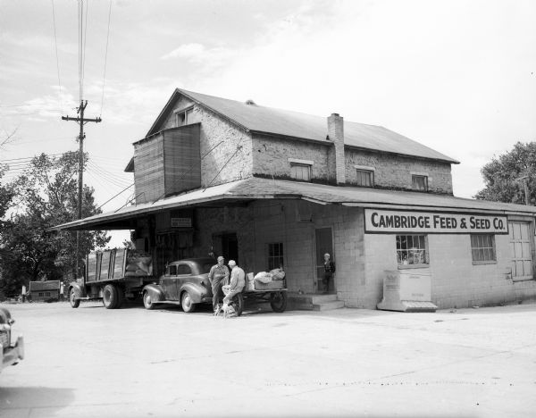 View across lot towards men, a boy, and a dog standing beneath an overhang outside the Cambridge Feed and Seed Company. Parked nearby are a truck and an automobile.