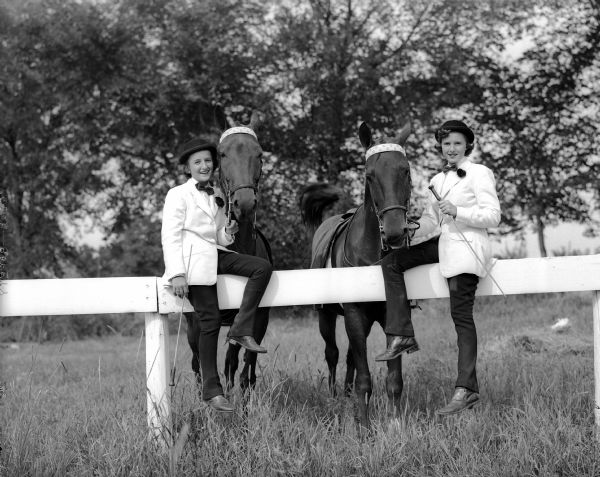 Two unidentified women in riding costume sit on a wooden fence while holding the reigns of their horses.