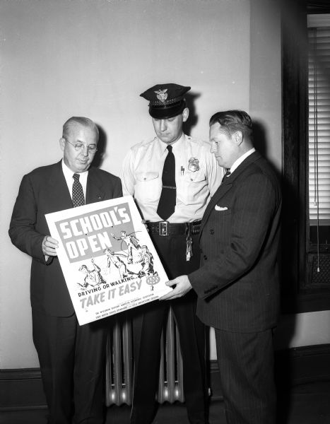 The American Automobile Association sponsored a campaign for safety at the fall opening of the schools with posters announcing "School's Open," displayed here by a police officer and two representatives of the AAA.