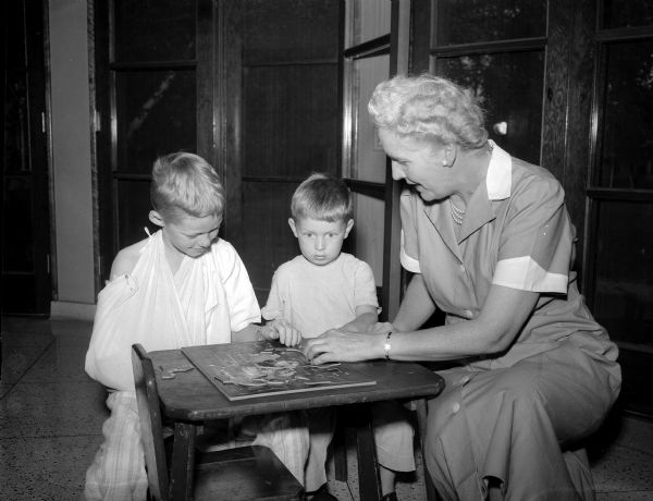 Florence Crowley, an active volunteer at St. Mary's Hospital, working on a jigsaw puzzle with Ronald Paar, age 9 of Cross Plains, and Kenneth Krall, age 4 of Madison. The hours of a hospital stay for children are long without the assistance of a pleasant lady in a peach uniform reading a story or offering another diversion.