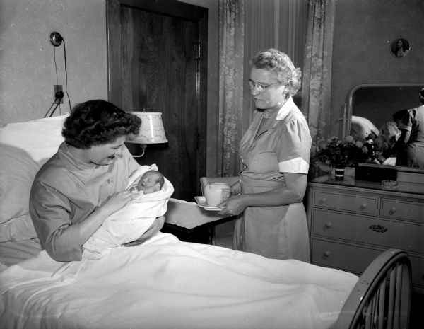 Volunteers supplement the nursing staff in many ways to accomplish the many tasks in a modern hospital. St. Mary's hospital is fortunate in having an organized women's auxiliary to aid the regular staff. Mrs. Thomas (Elizabeth) O'Neill brings a drink of water to Mrs. Russell A. (Laverne) Hart, pausing to admire Bradford Thomas Hart, born September 15, two months prematurely. Elizabeth O'Neill is secretary of the St. Mary's auxiliary.