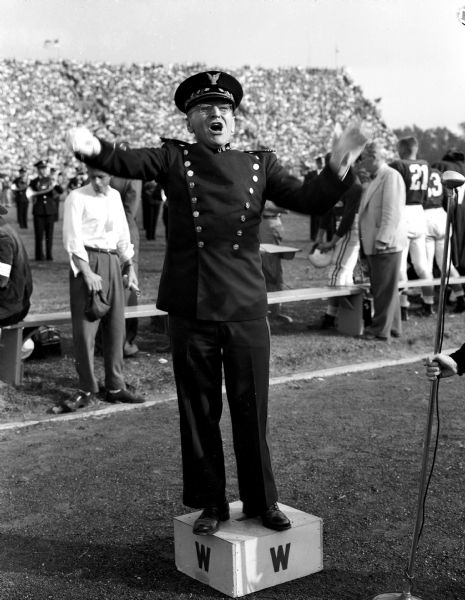 U.W. band director Ray Dvorak directs after returning from a two year battle with injuries suffered in a train wreck. He is shown in uniform directing the crowd of 45,000 in singing "Varsity" with his artificial right hand and arm.