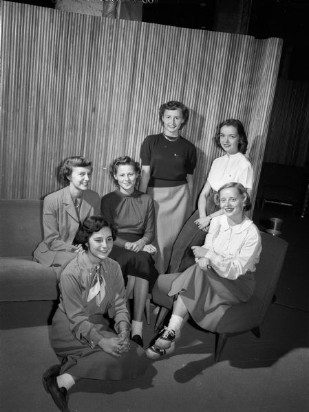 Four of the sixteen "ideal sorority girls" chosen by their sororities to represent them at the Wisconsin Panhellenic Ball, and two members of the planning committee. Seated on the floor is Nadine, Schuster, Madison, member of Alpha Epsilon and publicity chairman for the ball, and seated at extreme right is Winnie Weix, Oconomowoc, Kappa Kappa Gamma, general chairman of the dance. Left to right are four of the "ideal" girls, all from Madison, left to right: Jane Lewis, Alpha Xi Delta; Mary McCabe, Sigma Kappa; Deane Hanson, Alpha Gamma Delta, and Mona King, Theta Phi Alpha.