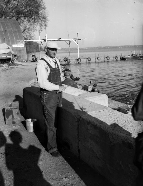 An unidentified man fishing along the shore of Lake Monona behind Fauerbach's Brewery, located at 651-3 Williamson Street. Four Lakes Boat Company, 425 South Blount Street, is in the background.