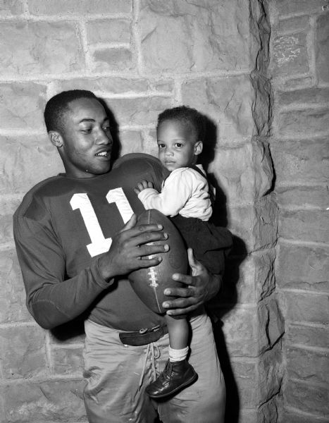 Eddie Withers, #11 on the University of Wisconsin football team, is shown with his 18 month old son, Eddie Withers III, after the Wisconsin team beat Iowa in a football game. Eddie also served in World War II, while he was a high school student. He was the first black football player to earn the rank of All-American in Wisconsin. He was posthumously inducted into the Madison Sports Hall of Fame in 1977.