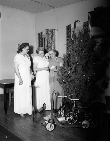 Three members of the Women of the Moose examine gifts to be sent to the children at Mooseheart by the Madison chapter while standing beside a Christmas tree. Left to right are: Mrs. Hazel Lerwick, Mrs. Edith Meier, and Mrs. Christine Deans.