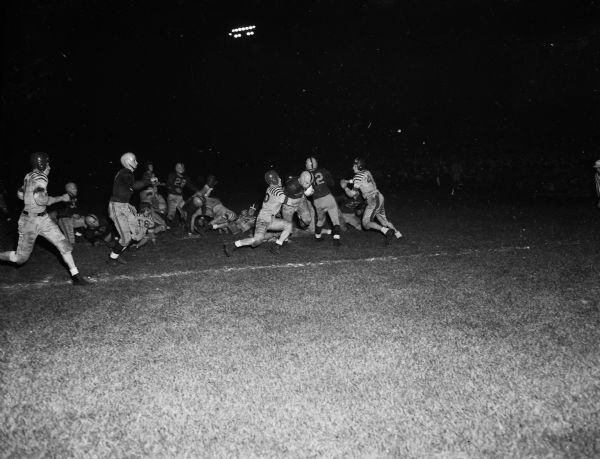 Pictured is the play that started Madison East High School on the way to its victory over previously-undefeated Madison West High School in their Big-Eight and intra-city high school football game. Ronnie Schara (#40), East, is shown carrying the ball for the final yard of the first touchdown. He is being tackled from the left by Jim Class (#30). Dick Falch (#52), East, is shown to Schara's left and Bill Mansfield (#4?), West, is coming up to meet the play. Glenn Barry (#44), West, is on the ground to the left of Class and Dale Pollock (#27), East, may be seen on his knees below Mansfield's arms.