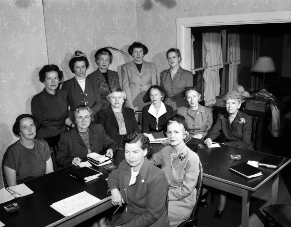 Group portrait of the Visiting Nurse Service Board of Directors. The function of the visiting nurses is to provide bedside care when needed in Madison. Standing in the background from left to right are: Janet Ela, Grace Frautschi, Madeline Hefty, Jean Antonius, and Mary O. Johnson; seated around the outer edge of the table from left to right are: Jane Rikkers, Mrs. Elbert H. Carpenter, Ann Schmich, Virginia Sprague, Esther Hermsen, and Mrs. Gilbert H. Doane; immediated foreground (left to right) Marian Stevens and Mrs. Francis F. Bowman Jr.