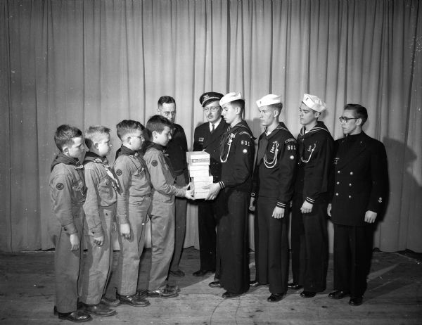 As part of Madison's civil defense plans, Boy Scout troops are being mobilized to distribute diabetic kits. Members of Scout Troop Number 2 are shown receiving kits from members of Sea Scout Ship "Wisconsin" Number 501.  Left to right are Gerald Rose, John Schwab, Bill Ketchum, John Buck and Scoutmaster John Thompson, all of Troop 2; and Skipper George T. Howe, John Forrest, Fritz Turner, Pat Kleinheinz and Robert Grant, all of Ship 501.